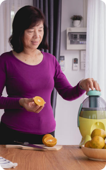 A senior woman stands in a kitchen usings a juicing machine to make fresh orange juice.