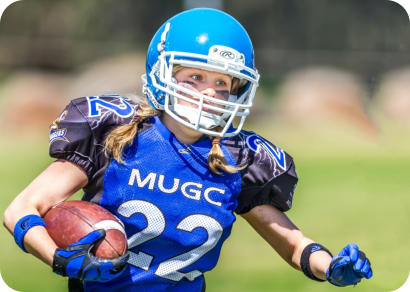 Closeup of a school-aged girl playing American football. She is wearing a blue uniform and helment, and is running with the football tucked under one arm.