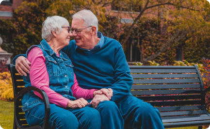 A senior couple sits together outside on a bench on a nice autumn day. The man has his arm around the woman and they looking at each other and smiling.